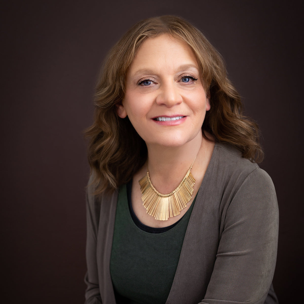 A smiling woman with shoulder-length wavy brown hair, wearing a dark green top, a taupe-brown cardigan, and a prominent gold statement necklace, set against a solid dark brown studio background.